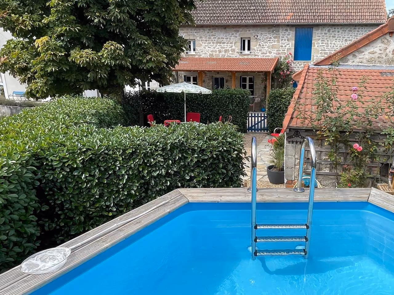 Houten bovengronds zwembad met roestvrijstalen ladder, met achter de haag het stenen huis en een parasol op het terras