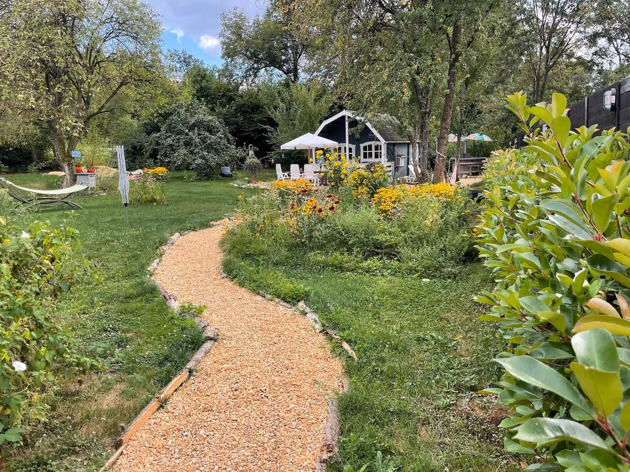 Grindpad door de tuin naar het donkergroene houten chalet, met bloemenborders, hangmat en een parasol